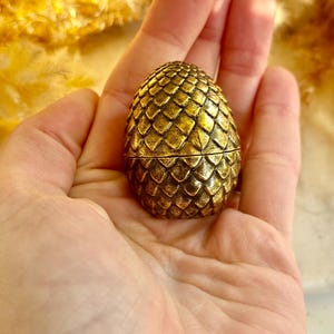 May include: A small, golden egg-shaped container with a textured, scale-like pattern. The egg is held in an open hand, showcasing its intricate design and metallic finish. The container appears to be made of metal and has a hinged opening.