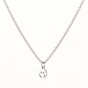 May include: A silver-toned metal necklace featuring a delicate cable chain and a small, circular pendant. The pendant is engraved with a simple smiley face design, consisting of two small circular eyes and a curved line representing a smiling mouth.