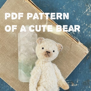 May include: PDF pattern for a cute white teddy bear, standing on a brown book with a green crystal. The bear is 8-9 cm tall. The text "PDF PATTERN OF A CUTE BEAR" is at the top of the image. The text "BEAR HEIGHT 8-9 CM" is at the bottom of the image.