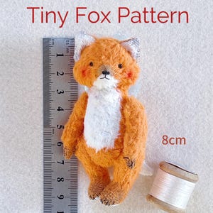以下が含まれることがあります： 定規と白い糸の糸巻きが置かれた横に、高さ8cmのオレンジと白の小さなキツネのぬいぐるみが置かれています。キツネは白いお腹と黒い鼻をしています。