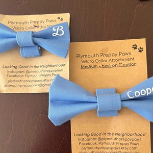 May include: Two light blue fabric bow ties for pet collars, each attached to a tan cardboard display card. One bow tie features the white letter B, while the other displays the white name Cooper. Both cards include text about Plymouth Preppy Paws.