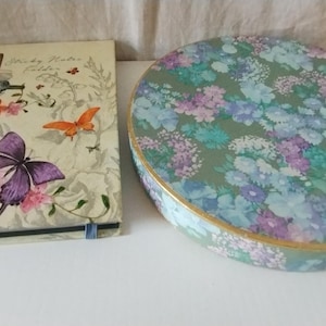 May include: A floral patterned notebook with a butterfly and hummingbird design. The text on the cover reads "Shady Meadow Garden". A round floral patterned box with a gold trim.