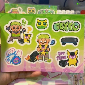 May include: A sheet of stickers featuring cartoon characters and the word "GEKKO" in green. The stickers include a skateboard, a blue and pink explosion, and a yellow creature with a purple mask. The artist's name, "@meteorite_studios," is also visible.