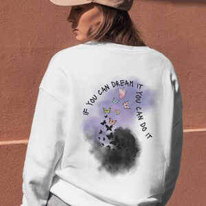 Könnte beinhalten: Weißes Sweatshirt mit Schmetterlingsdesign und dem Text "If you can dream it, you can do it." Das Design zeigt bunte Schmetterlinge, die aus einer dunklen Wolke vor einem lila Hintergrund auftauchen.