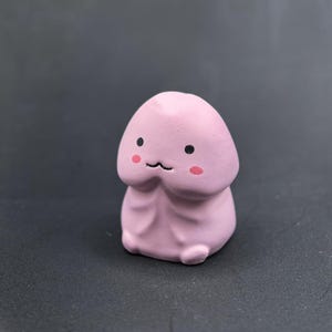 Puede incluir: Una figura rosa de estilo caricaturesco con forma de falo. La figura tiene una cara sonriente con ojos negros y marcas rosadas en las mejillas. El fondo es de color gris oscuro sólido.