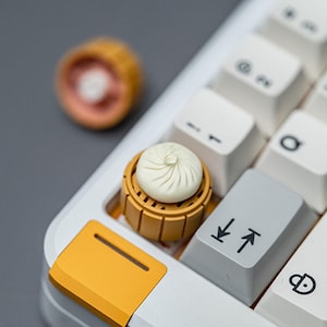 May include: A white and yellow mechanical keyboard with a custom keycap shaped like a dumpling in a bamboo steamer. The keycap is white and the steamer is yellow. The keyboard keys are white and gray.
