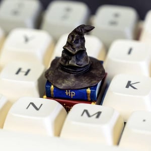 May include: A miniature Sorting Hat figurine atop two small books with gold trim and the letters "Hp" on the blue book. The figurine rests on a white keyboard, with the keys "N", "M", "H", "I", and "K" visible.