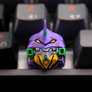 May include: A custom keycap featuring a purple, yellow, green, and gray robot head design. The keycap is a detailed, stylized representation of a character, likely for a mechanical keyboard.