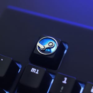May include: A close-up shot of a black keyboard with a custom keycap displaying the Steam logo. The keycap has a silver border and a blue illuminated centre. The keyboard keys are black with white lettering.