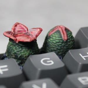 May include: Two small, green, textured sculptures with red accents, resembling alien plant life, rest on a computer keyboard. The keyboard keys are gray, with the letter "G" visible on one key. The sculptures have a detailed, organic appearance.