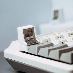 Puede incluir: Un pequeño juguete de ordenador retro blanco con una pantalla que muestra un gráfico, colocado en un teclado blanco y gris. La pantalla muestra un gráfico pixelado. Las teclas del teclado son blancas y grises.