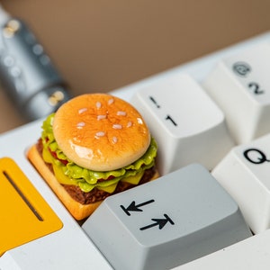 Pode incluir: Um mini-hambúrguer detalhado, com sementes de sésamo no pão, alface, tomate e queijo, repousa sobre um teclado de computador. O hambúrguer tem um acabamento brilhante. As teclas do teclado são brancas e cinzentas.