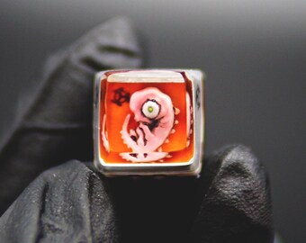 May include: A square, clear resin keycap with a pink, stylized embryo design set against an orange background. The keycap has a silver-colored metal frame. The embryo has a single green eye.