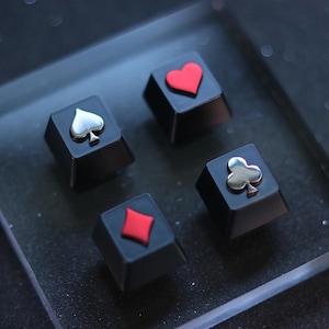 Peut inclure: Quatre touches de clavier noires avec des symboles de jeu de cartes : pique, cœur, carreau et trèfle. Les symboles de pique et de trèfle sont argentés, tandis que le cœur et le carreau sont rouges. Les touches sont dans un récipient en plastique transparent.