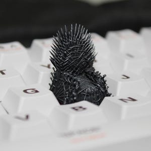 Könnte beinhalten: Eine detaillierte, kleine schwarze Iron Throne-Figur sitzt auf einer weißen Computertastatur. Der Miniaturthron hat eine stachelige Rückenlehne und Armlehnen, die mit den glatten, weißen Tastenkappen kontrastieren. Die Tastaturtasten sind mit schwarzen Buchstaben beschriftet.