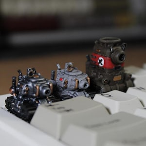 May include: Three miniature toy tanks in shades of grey, black, and brown, with intricate details. One tank has a red stripe and a white symbol. The tanks are positioned on a white keyboard.