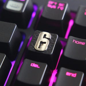 May include: A close-up of a black mechanical keyboard with illuminated keys. The keycap in focus features a metallic silver "6" with a gun symbol. Other keys include "home" and "del" with pink backlighting, suggesting a gaming setup.