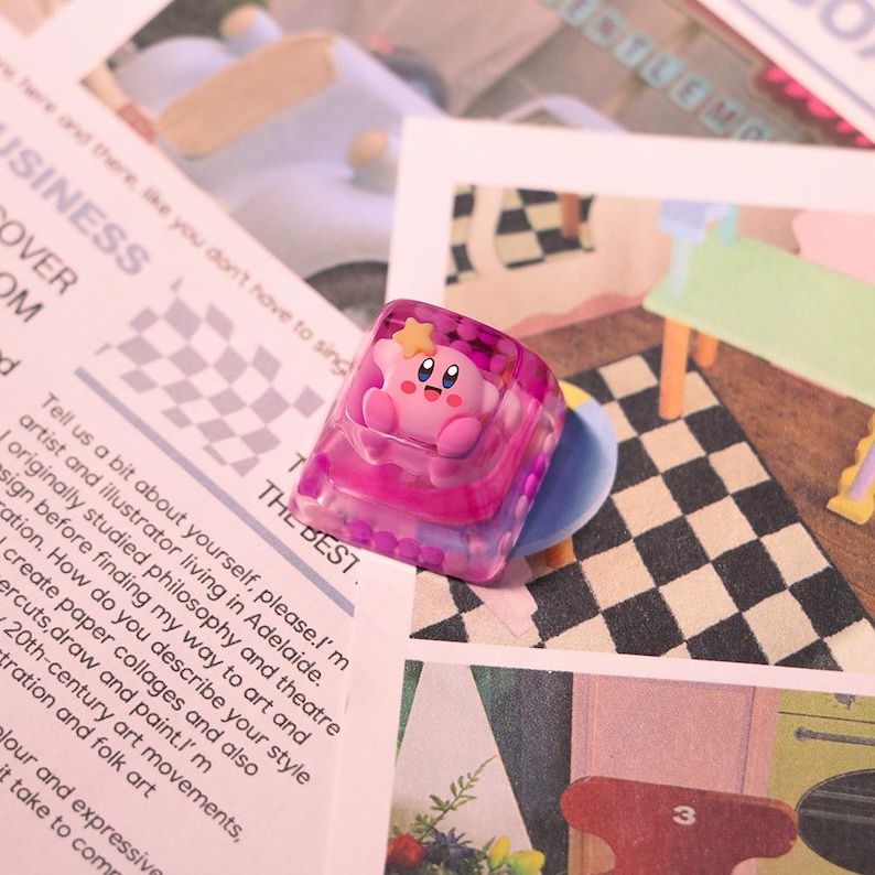 May include: A pink and clear resin keycap featuring a cartoon character with a star on its head. The keycap has a square shape with rounded edges and is set against a background of printed paper with text and a checkered pattern.