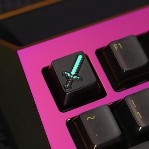 May include: Close-up of a black keyboard key with a pixelated sword design in teal and gold. The key is surrounded by other black keys and a bright pink and black keyboard base. The F1 key is also visible.