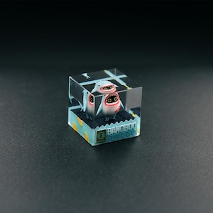 Może przedstawiać: Przezroczysty, sześcienny element artystyczny z żywicy, przedstawiający postać rekina z kreskówki. Rekiny są biało-czerwone z czarnymi oczami, umieszczone na niebieskiej platformie z napisem "BANGBOO".