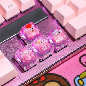 May include: Close-up of a pink mechanical keyboard with custom keycaps. The keycaps feature a cartoon character design with pink and white colors. The keyboard has a pink base and pink keycaps.