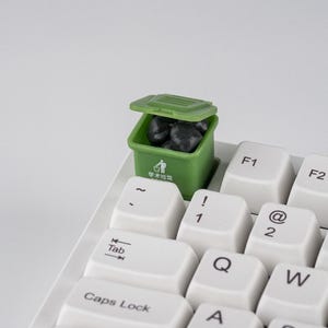 Op de afbeelding: Een kleine groene vuilnisbak, gevuld met zwarte voorwerpen, staat op een wit toetsenbord. De bak heeft een deksel en Chinese tekst. Witte toetsen met zwarte letters zijn zichtbaar.
