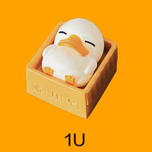 May include: A white duck figurine with a yellow beak and feet rests in a light brown box. The duck has closed eyes, suggesting it is sleeping. The box has decorative elements on the front. The number "1U" is at the bottom.