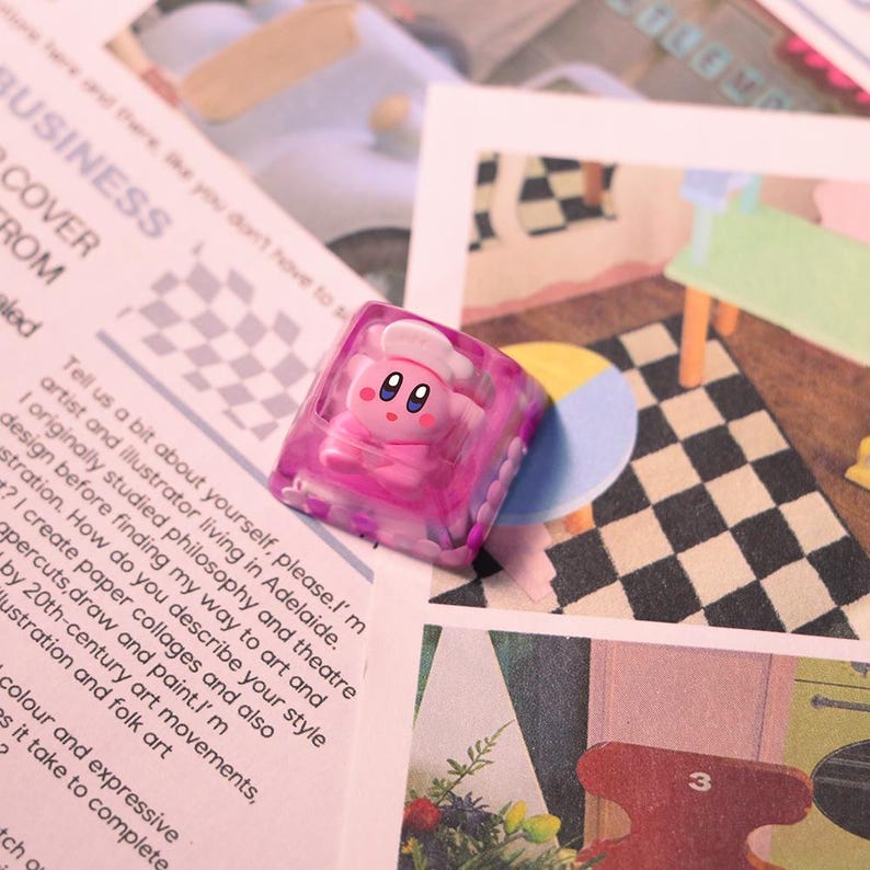 May include: A pink and clear resin keycap featuring a cartoon character with a chef's hat. The keycap is set against a backdrop of printed paper and illustrations, with a focus on the keycap's design and color.