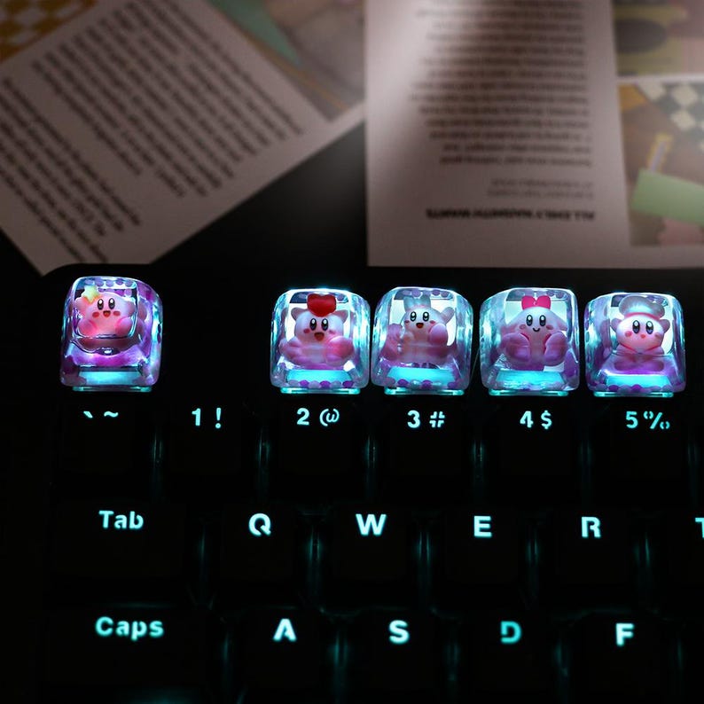 May include: A set of five custom keyboard keycaps featuring a pink cartoon character inside a clear, cube-shaped design. The keycaps are illuminated with a blue glow, and are placed on a black keyboard. The keys are labeled with numbers and symbols.