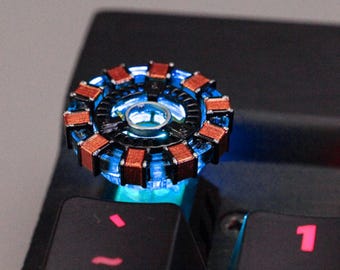 May include: A glowing, circular device with a blue center and a ring of copper-colored rectangular components. The device is placed on a black keyboard, with the keys visible below. The device emits a blue light.