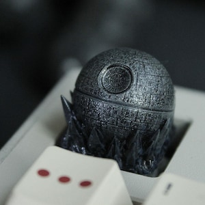 Puede incluir: Una detallada tecla Death Star metálica, un diseño esférico con intrincados detalles superficiales, se encuentra sobre un teclado de ordenador. La tecla es gris con una base oscura, y las teclas del teclado son blancas.