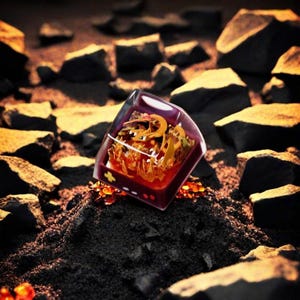 May include: A custom keycap with a transparent, square design. Inside, a fiery orange and yellow resin art piece is visible, with a golden-colored dragon-like figure. The keycap sits on a bed of dark, textured material, with dark rocks in the background.