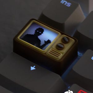 May include: A custom keycap shaped like a vintage television set. The screen displays a silhouette of a figure with glowing eyes. The keycap is brown and beige, with two knobs and a speaker grill. It is set on a gray keyboard.