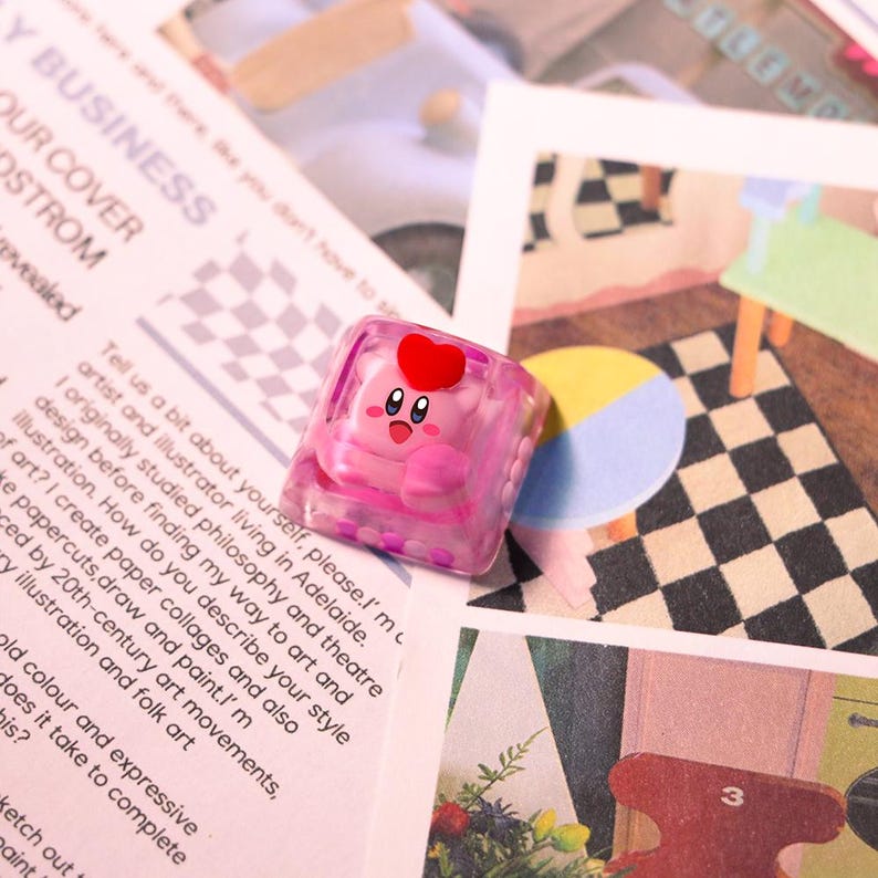 May include: A pink, square-shaped keycap featuring a cartoon character with a red heart on its head. The keycap is encased in a clear, pink-tinted resin. The background includes printed paper with text and colorful illustrations.