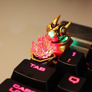 May include: A custom keyboard keycap with a gold figurine and a pink crystal, set on a black keyboard. The keycap has green accents and pink illuminated lettering on the keyboard.