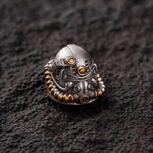 Peut inclure: Un casque miniature argent&eacute; avec des d&eacute;tails dor&eacute;s. Le casque pr&eacute;sente un design m&eacute;canique complexe, avec un &eacute;l&eacute;ment de cou c&ocirc;tel&eacute; et des &eacute;l&eacute;ments circulaires. Il est pos&eacute; sur un fond sombre et textur&eacute;, cr&eacute;ant un contraste saisissant.