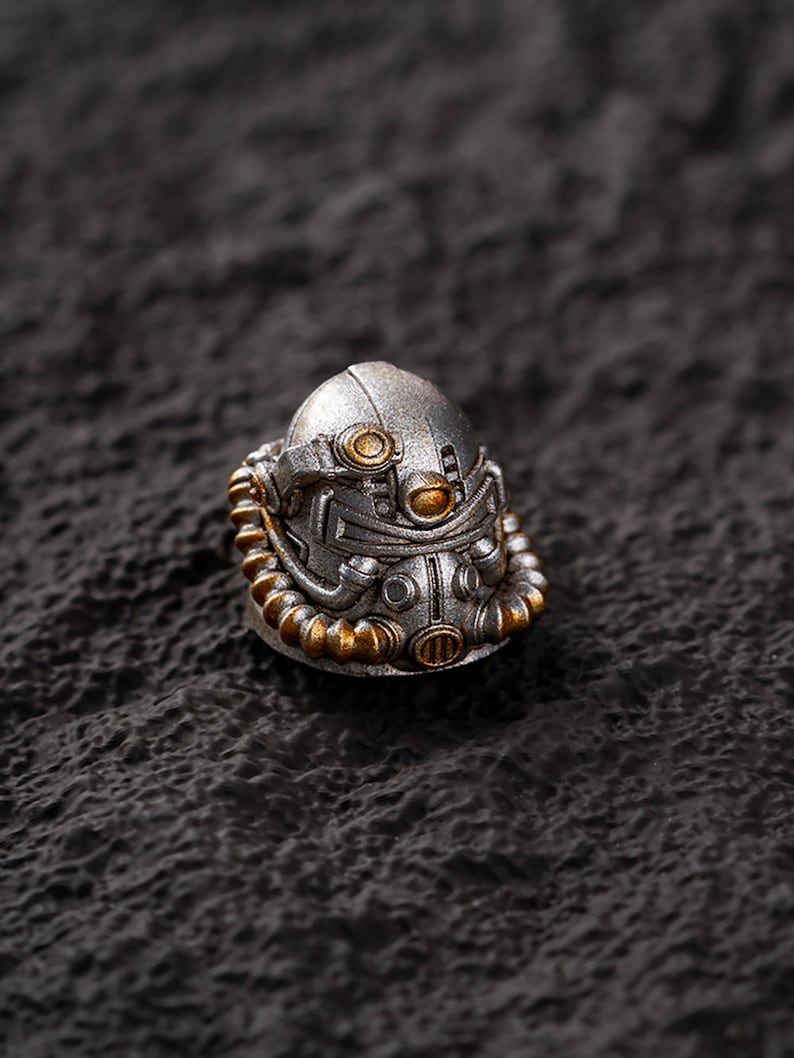 Peut inclure: Un casque miniature argent&eacute; avec des d&eacute;tails dor&eacute;s. Le casque pr&eacute;sente un design m&eacute;canique complexe, avec un &eacute;l&eacute;ment de cou c&ocirc;tel&eacute; et des &eacute;l&eacute;ments circulaires. Il est pos&eacute; sur un fond sombre et textur&eacute;, cr&eacute;ant un contraste saisissant.