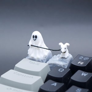 May include: A novelty keyboard accessory showing a white ghost figure walking a small white dog on a leash. The ghost has a smiling face. The figures are positioned on grey keycaps, with other black and grey keys visible.