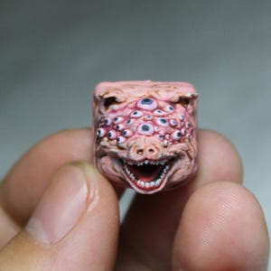 May include: A detailed, custom-made artisan keycap featuring a grotesque, fleshy design. The keycap is a light pink color and covered in multiple blue-eyed sockets. The bottom of the keycap has a wide, toothy grin.