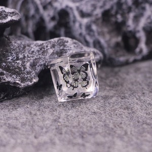 May include: A clear, square-shaped decorative object with black butterfly designs. The butterflies are depicted in various sizes and positions, adding a whimsical touch. The object is set against a gray background.