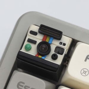 May include: A white and black camera-shaped keycap with a rainbow stripe detail. The keycap has a green button and a black lens. It is placed on a gray keyboard.
