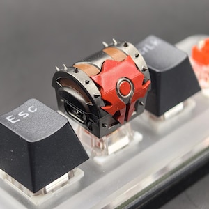 May include: A custom mechanical keyboard keycap shaped like a treasure chest. The keycap is primarily black with brown and red accents, and features a detailed design with spikes and a circular emblem. The keycap is placed between other black keys.