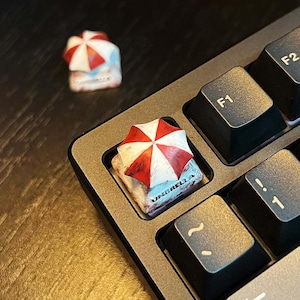 May include: A custom keyboard keycap featuring a miniature red and white umbrella design. The keycap is square and sits on a gray keyboard, with the word "UMBRELLA" visible on the keycap.