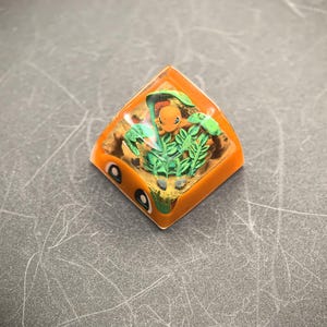May include: An orange, diamond-shaped artisan keycap featuring a small orange creature surrounded by green leaves and foliage. The keycap has a clear resin top, and the creature is set against a sandy background. The keycap has two white and black eye-like details.
