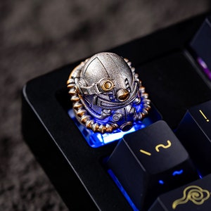 Peut inclure: Une touche artisanale argent&eacute;e en forme de casque futuriste, mont&eacute;e sur un clavier m&eacute;canique noir. La touche pr&eacute;sente des d&eacute;tails dor&eacute;s et est &eacute;clair&eacute;e par une lumi&egrave;re bleue. Les touches du clavier ont des symboles dor&eacute;s.