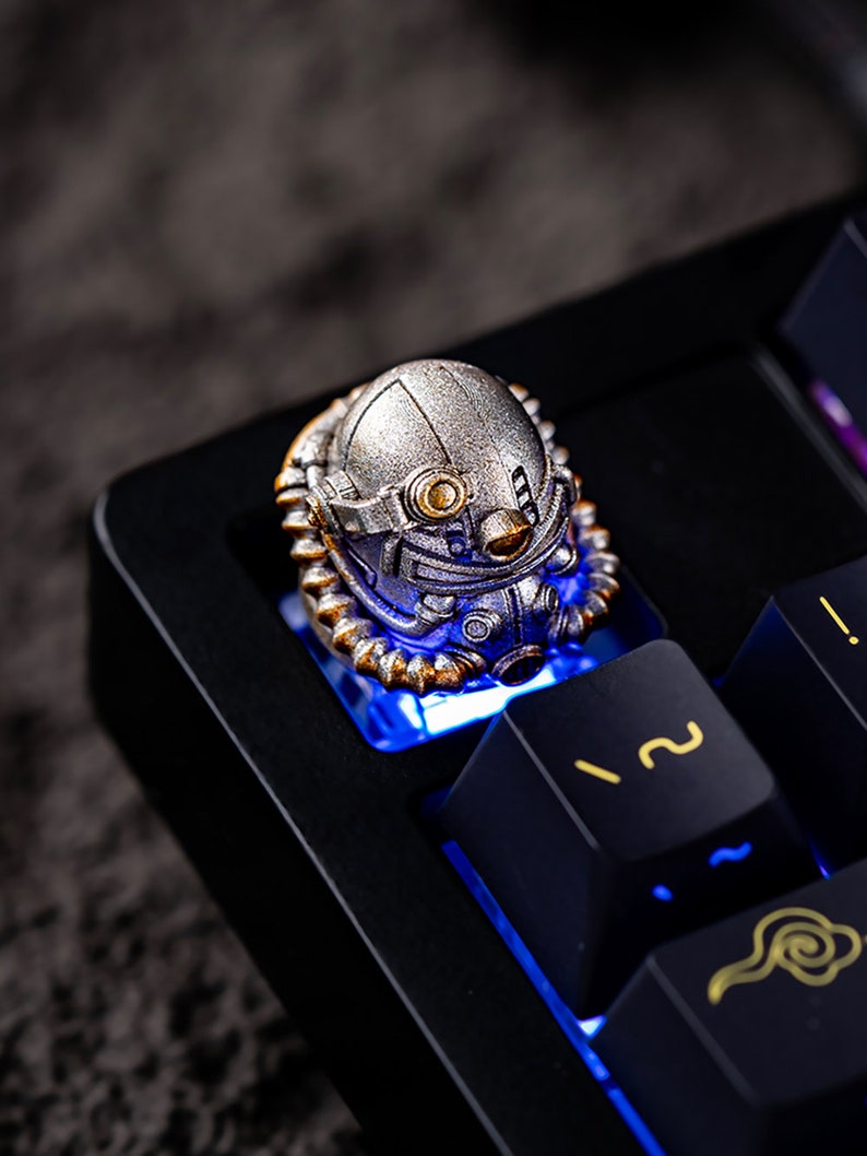 Peut inclure: Une touche artisanale argent&eacute;e en forme de casque futuriste, mont&eacute;e sur un clavier m&eacute;canique noir. La touche pr&eacute;sente des d&eacute;tails dor&eacute;s et est &eacute;clair&eacute;e par une lumi&egrave;re bleue. Les touches du clavier ont des symboles dor&eacute;s.