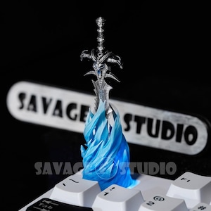 May include: A detailed silver-colored sword figurine with blue accents, seemingly emerging from a blue flame base. The word "SAVAGE STUDIO" is visible in the background. The figurine is placed on a white keyboard.