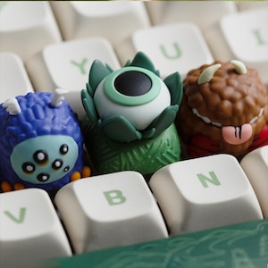 Op de afbeelding: Close-up van drie kleurrijke keycaps met monsterthema's op een toetsenbord. De keycaps tonen een blauw monster met meerdere ogen, een groen oogvormig monster en een bruin monster met hoorns en een tong. De toetsenbordtoetsen zijn wit.