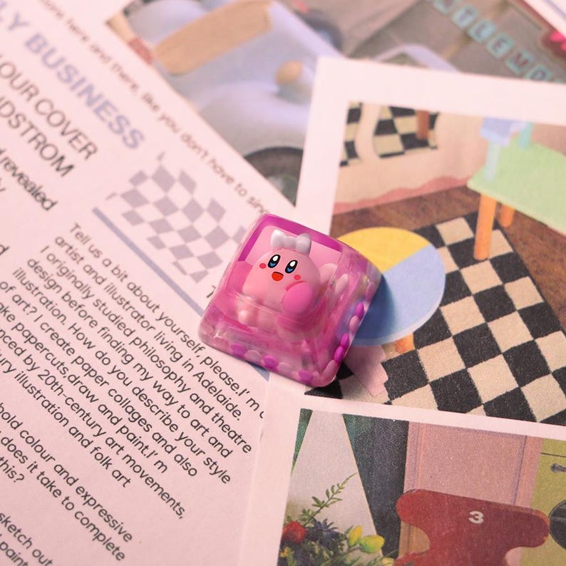 May include: A pink and clear resin keycap featuring a cartoon character with a white bow. The keycap is set against a backdrop of printed paper with text and illustrations, including a checkered pattern.