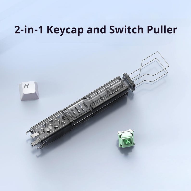 May include: A 2-in-1 keycap and switch puller tool. The tool is dark gray and clear, with a wire keycap puller and a switch puller. A white keycap with the letter "H" is next to the tool, along with a green switch.